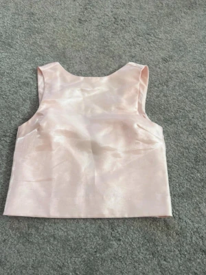 Ljusrosa satinväst med rosett - Ljusrosa väst i satin med v-ringning och en dekorativ rosett framtill. Västen har en rak och kort passform, vilket ger en stilren och elegant look. Materialet är glansigt och mjukt, perfekt för att addera lite extra till din outfit.