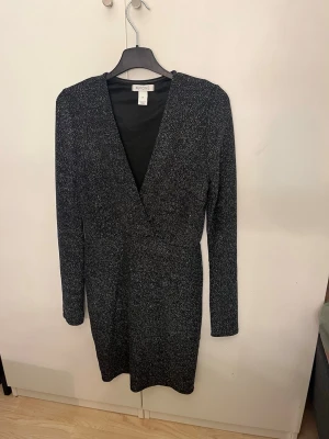 Glittrig svart omlottklänning NYONE - Snygg svart kortklänning från NLYONE med glittrigt tyg och omlott-design. Klänningen har lång ärm och djup v-ringning, perfekt för dig som vill sticka ut. Materialet är mjukt och följsamt, vilket ger en bekväm passform.