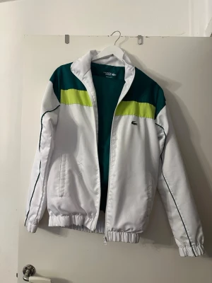 Lacoste vit och grön träningsdress - Säljer en retro träningsdress från Lacoste i vitt, grönt och limegult. Jackan har dragkedja, ståkrage och elastiska muddar. Byxorna är gröna med vita detaljer och resår i benslut. Perfekt för sport eller chill. Klassisk krokodil-logga på bröstet.