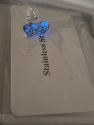 Hjärtformade glow örhängen i stål - Säljer ett par snygga hjärtformade örhängen i rostfritt stål. De lyser blått i mörker efter att ha varit en liten stund i dagsljus. Örhängena har ett öppet hjärtmönster och hänger på krokar. Perfekta för dig som gillar stilrena och romantiska accessoarer.