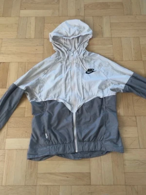 Nike vindjacka i grått och vitt - Nike vindjacka med huva i tvåfärgad design, ljusgrå upptill och mörkare grå nedtill. Jackan har dragkedja framtill, elastisk nederkant och fickor med dragkedja. Klassisk Nike-logga på bröstet. Perfekt för en sportig och avslappnad stil.