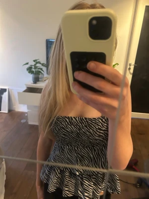 Zebramönstrad bandeau topp - Säljer en zebramönstrad bandeau topp med volang nedtill. Toppens smockade överdel ger en tight passform och den är ärmlös med axelband. Perfekt för dig som vill sticka ut med ett djurmönster och en cool look.