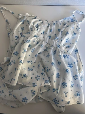 Vit blommig sommartopp - Söt vit topp med blå blommor och smala axelband. Toppen har knappar framtill och ett lätt, luftigt tyg som passar perfekt för varma dagar