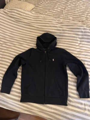 Mörkblå hoodie från Polo Ralph Lauren - Snygg mörkblå hoodie med dragkedja från Polo Ralph Lauren. Klassisk design med huva och dragsko, två fickor framtill och den ikoniska vita logon broderad på bröstet. Perfekt för en chill och stilren look. Materialet är mjukt och bekvämt, troligen bomullsblandning.