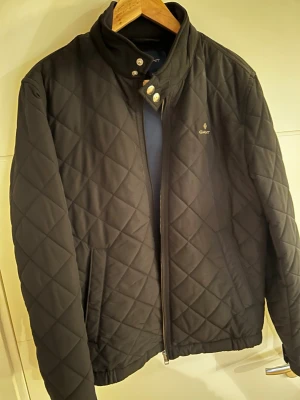 Svart quiltad jacka  - Snygg svart quiltad jacka från Gant med hög krage och dragkedja framtill. Jackan har diskret logga på bröstet, tryckknappar vid halsen och ett klassiskt rutmönster. Perfekt för höst och vår när du vill ha en stilren look.