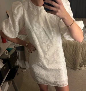 Studentklänning - Säljer en vit kortklänning med puffiga ärmar och diskret blommönster i tyget. Klänningen har rund halsringning och är tillverkad i ett strukturerat material. Klänningen är uppsydd så det är lite kortare än när jag köpte den, men väl uppsydd. 