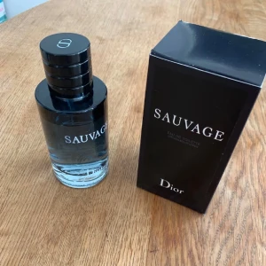 Dior Sauvage Eau de Toilette - Dior Sauvage Eau de Toilette med en elegant svart och transparent glasflaska. Flaskan har en rund form och ett stilrent svart lock med Dior-loggan. Förpackningen är svart med vit text och utstrålar lyxig och modern känsla.
