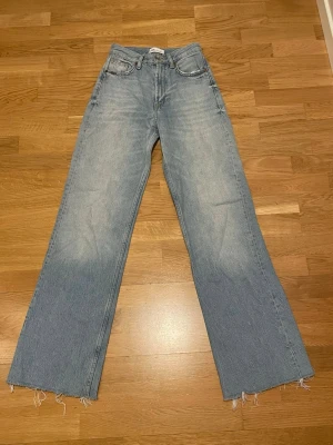 Ljusblå raka jeans med rå kant - Säljer ett par ljusblå jeans med rak passform och rå, fransig kant nedtill. Jeansen har klassisk femficksdesign och hög midja. Perfekta för en avslappnad och trendig look. Materialet är denim i bomull.