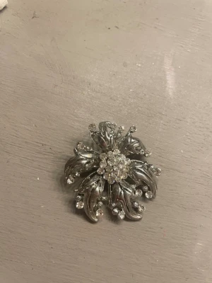 Silvrig vintage brosch med strass - Unik silvrig brosch i form av en blomma med gnistrande klara strasstenar i mitten och längs kronbladen. Broschen har en elegant retrostil och är tillverkad i metall med dekorativa detaljer. Perfekt för att ge din outfit en extra touch av glamour.