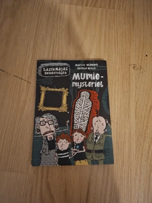 LasseMajas Detektivbyrå: Mumiemysteriet - En spännande och rolig bok ur serien LasseMajas Detektivbyrå, skriven av Martin Widmark och illustrerad av Helena Willis. Omslaget är färgglatt med tecknade figurer och en mumie i centrum. Perfekt för unga läsare som gillar mysterier.