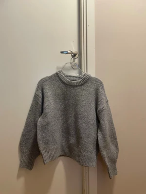 Grå stickad tröja med rund hals - En enkel och stilren grå stickad tröja med rund halsringning och ribbade muddar vid ärmslut och nederkant. Tröjan har en avslappnad passform och är perfekt för lager på lager under kyliga dagar. Materialet ser ut att vara mjukt och bekvämt. Vid halsringningen finns små få rosa prickar kan kanske gå bort vid tvätt vet ej