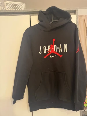 Svart Jordan hoodie med röd logga - Snygg svart hoodie från Jordan med stor röd Jumpman-logga och vit text på bröstet. Har även en mindre röd logga på ärmen och klassisk känguruficka framtill. Tillverkad i mjukt material och har en stor huva för extra komfort.