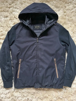 Marinblå vårjacka med huva - Snygg marinblå vindjacka från Massimo Dutti med huva och dragkedja framtill. Jackan har två fickor med kontrasterande dragkedjor och ribbade muddar vid ärmsluten. Materialet är en mix av slätt tyg och mjukare tyg på ärmarna för en cool look passar perfekt till våren.