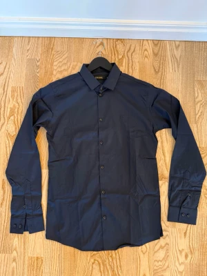 Mörkblå skjorta - Stilren mörkblå skjorta med klassisk krage och knäppning framtill. Skjortan har långa ärmar med manschett och är tillverkad i ett slätt bomullstyg. Perfekt för en clean och enkel look. Köpt i Spanien, och är använd ett fåtal gånger.