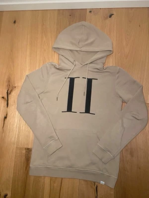 Les Deux Hoodie  - Säljer en beige hoodie med stor svart II-logga på bröstet. Hoodien har huva med dragsnören, ribbade muddar och en klassisk magficka. Materialet är mjukt och bekvämt, perfekt för en avslappnad stil.