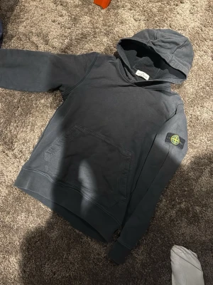 Mörkgrå hoodie från Stone Island - Snygg mörkgrå hoodie från Stone Island med klassisk patch på vänster ärm. Hoodien har en stor känguruficka framtill och en skön huva. Perfekt för en avslappnad streetwear-look.