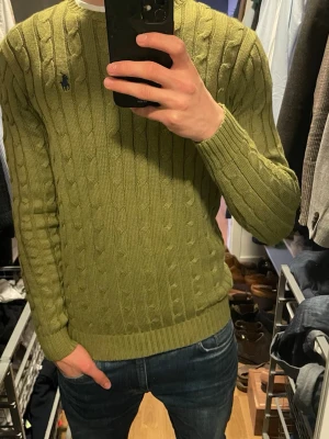 Olivgrön kabelstickad tröja Polo Ralph Lauren - Kabelstickad olivgrön tröja från Polo Ralph Lauren med rund halsringning och broderad logga på bröstet. Tröjan har långa ärmar och ribbade muddar vid ärmslut och nederkant. Perfekt för dig som gillar klassisk och stilren design.