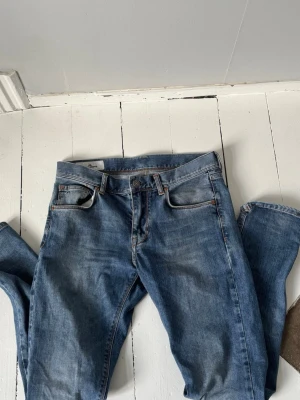 Jeans från J.Lindeberg - Säljer ett par blå jeans med rak passform och klassisk femficksdesign. Jeansen har en snygg tvättad finish och detaljer som kontrastsömmar och läderpatch bak i midjan. Använda ett fåtal gånger, pris går att diskutera!
