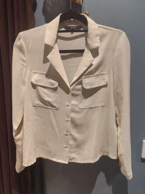 Beige skjorta  - Stilren beige skjorta med klassisk krage, två bröstfickor och knappar framtill. Skjortan har lång ärm och är tillverkad i ett mjukt, lätt material som ger en avslappnad men ändå snygg look. Perfekt att matcha med jeans eller kjol.