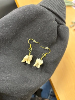 Unika örhängen med tandhängen - Säljer ett par riktigt unika örhängen med guldfärgade krokar och hängande tandformade detaljer i benvit färg. Perfekt för dig som gillar statement-smycken och vill sticka ut med något annorlunda och edgy.