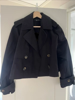 Kort svart trenchcoat från Gina Tricot - Snygg kort svart trenchcoat från Gina Tricot med dubbelknäppning, bred krage och dekorativa axelklaffar. Jackan har stora knappar och justerbara ärmslut med spännen. Perfekt för dig som vill ha en trendig och stilren look.