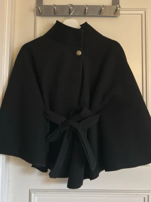 Missäy poncho - Stilren svart cape med hög krage och dekorativ knapp framtill. Capen har ett brett knytband i midjan som ger en snygg siluett. Materialet ser ut att vara mjukt och lite tjockare, perfekt för kyligare dagar. Enkel men elegant design utan synligt märke.