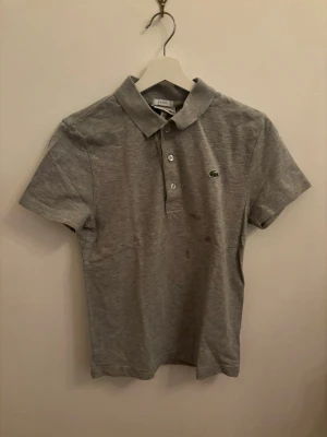 Grå pikétröja från Lacoste Sport - (Se bild för defekt) har ej testat att få bort. Klassisk grå pikétröja från Lacoste Sport i slim fit-modell. Tröjan har korta ärmar, krage och knappar framtill samt den ikoniska gröna krokodilloggan på bröstet. Perfekt till jeans eller shorts för en clean och sportig look.
