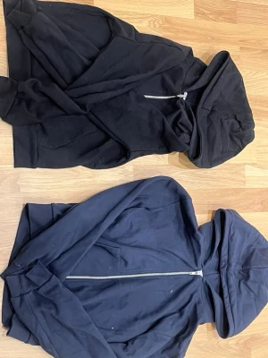Två ziphoodies i svart och mörkblå - Säljer två stilrena hoodies med dragkedja, en i svart och en i mörkblå. Båda har huva och är tillverkade i mjukt bomullsliknande material. Perfekta för en avslappnad och enkel look. Inga synliga loggor eller tryck.