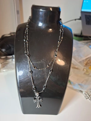 Dubbelradigt halsband med korshänge - Snyggt dubbelradigt halsband i silverfärgad metall med små svarta pärlor längs kedjan. Halsbandet har ett stort korshänge med detaljerade kanter och svarta stenar. Perfekt accessoar för en edgy och alternativ stil.