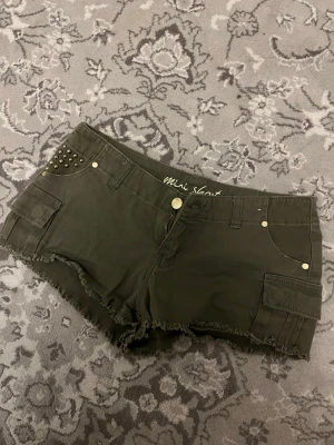 Mörkgröna shorts med nitar - Snygga mörkgröna shorts med rå kant och coola nitar på ena fickan. Shortsen har fickor både fram och på sidorna samt guldiga knappar och detaljer. Perfekta för en trendig och avslappnad stil.