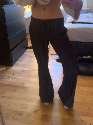 Mörkgrå randiga kostymbyxor - Säljer ett par mörkgrå kostymbyxor med tunna vita kritstrecksränder. Byxorna har en rak passform med lätt utsvängda ben och hög midja. Perfekta för en stilren och trendig look. Materialet känns mjukt och följsamt.