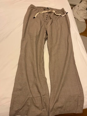 Beige rutiga linnebyxor från Lindex - Snygga beige linnebyxor från Lindex med rutigt mönster och avslappnad passform. Byxorna har resår i midjan och dragsko med dekorativ snörning samt knappar framtill. Perfekta för en chill och stilren look.