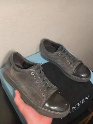 Gråa Lanvin sneakers med lackad tå - Säljer ett par gråa Lanvin sneakers i mocka med snygg lackad tå i svart. Skorna har klassisk låg profil, rundad tå och matchande grå snörning. Perfekta för dig som gillar stilrena och exklusiva detaljer. Kommer med originalkartong. Pris ej hugget i sten! Skriv vid minsta fundering!! 😃