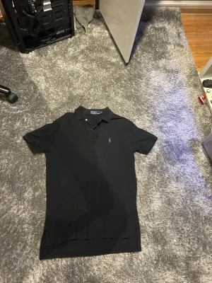  Ralph Lauren piké - Klassisk svart pikétröja från Ralph Lauren med lila broderad logga på bröstet. Tröjan har korta ärmar, krage och knappar framtill. Tillverkad i bomull för en bekväm känsla och snygg passform. Snygg inför våren och sommarn!