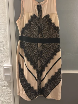 Beige klänning med svart spetsmönster - Snygg beige kortklänning med svart spetsmönster och markerad midja. Klänningen är ärmlös och har dragkedja bak. Perfekt för dig som gillar en elegant och modern look med coola detaljer. Från Elise Rayan med prislappen kvar. Står bara storlek 12, skulle tolkas som S 