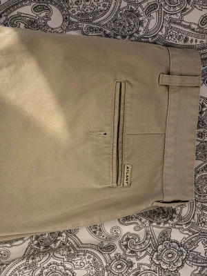 Beige chinos från Atlant - Snygga beige chinos från Atlant med klassisk rak passform och bälteshällor. Byxorna har en diskret bakficka med knapp och är tillverkade i ett mjukt bomullstyg som känns skönt mot huden. Perfekta för en clean och stilren look.