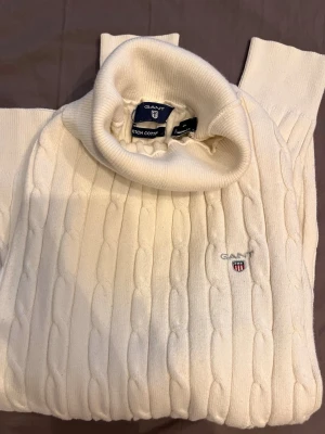 Vit polotröja från Gant - Kabelstickad vit polotröja från Gant i bomullsmix. Tröjan har hög polokrage, ribbade muddar och Gant-logga med sköld broderad på bröstet. Klassisk och stilren design. 