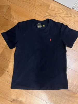 Svart t-shirt från Polo Ralph Lauren - Klassisk svart t-shirt från Polo Ralph Lauren med röd broderad logga på bröstet. T-shirten har rund halsringning och korta ärmar. Tillverkad i mjuk bomull för en skön och avslappnad känsla. Perfekt till jeans eller shorts.