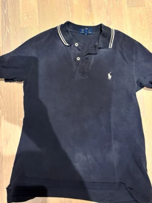 Marinblå pikétröja från Ralph Lauren - Klassisk marinblå pikétröja från Ralph Lauren med vit broderad logga på bröstet och kontrasterande vita detaljer vid kragen. Baksidan har texten 'POLO RALPH LAUREN' i vitt och rött längs kragen. Perfekt till en avslappnad men stilren look.