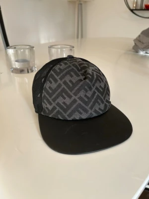 Svart Fendi keps med FF-mönster - Säljer en svart keps från Fendi med det klassiska FF-mönstret i grått över hela framsidan och mesh på sidorna. Skärmen är svart och justerbar rem baktill med guldfärgad Fendi-logga. Perfekt för dig som gillar exklusiva accessoarer och streetwear.