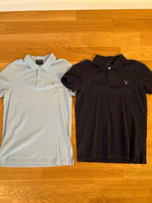 Pikétröjor - Säljer två klassiska blåa pikétröja från Gant med korta ärmar och krage. Tröjorna har knappar framtill och den ikoniska Gant-loggan broderad på bröstet. Perfekt för dig som gillar stilrena och tidlösa plagg.