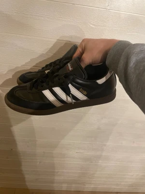 Adidas Samba svarta sneakers - Klassiska Adidas Samba sneakers i svart läder med vita detaljer och bruna gummisulor. Skorna har de ikoniska tre ränderna på sidan och guldfärgad Samba-text. Låg modell med snörning och retrovibe, perfekt för dig som gillar streetwear.
