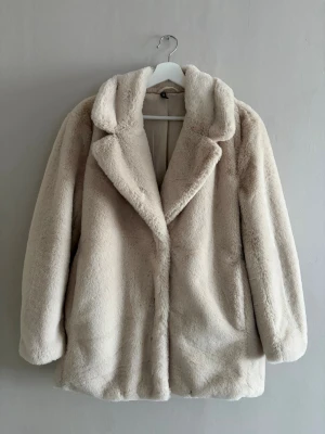Beige fuskpälsjacka med bred krage - Säljer en supermjuk beige fuskpälsjacka med bred krage och rak passform. Jackan har långa ärmar och stängs framtill. Perfekt för dig som vill ha en stilren och cozy look under kalla dagar.