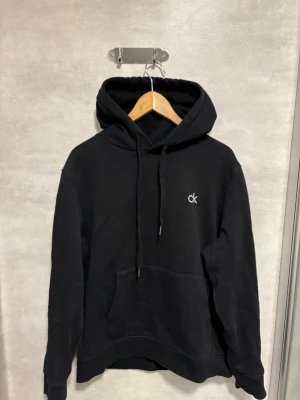 Svart hoodie från Calvin Klein - Svart hoodie från Calvin Klein med klassisk huva och dragsnören. Diskret 'ck'-logga broderad på bröstet och stor magficka framtill. Tillverkad i mjukt material som känns skönt mot huden. Perfekt för en avslappnad och stilren look.