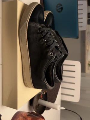 Svarta lanvins  - Svarta sneakers med klassisk design, rund tå och snörning. Ovandelen är i mocka och tåpartiet i skinn, vilket ger en snygg kontrast. Skorna har en vit platt sula som ger en sportig vibe. Perfekta för dig som gillar stilrena och enkla sneakers.