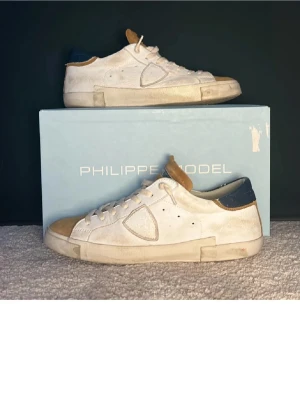 Vita Philippe Model sneakers låga - Snygga vita sneakers från Philippe Model med beige mockadetaljer på tån och plösen samt mörkblå häl. Skorna har klassisk låg silhuett, vita skosnören och en robust sula. Perfekta för dig som gillar stilrena och trendiga sneakers.