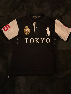 Ralph lauren piké Tokyo - Snygg svart t-shirt från Polo Ralph Lauren med vita ärmar och stora röda siffror 5 på ärmen och ryggen. Framtill står det TOKYO i vitt, samt har en broderad logga och emblem. Klassisk pikékrage och knappdetaljer framtill.