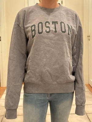 Grå sweatshirt med BOSTON-tryck - Säljer en grå sweatshirt med texten BOSTON i stora bokstäver över bröstet. Tröjan har rund halsringning, långa ärmar och ribbade muddar vid ärmslut och nederkant. Perfekt för en avslappnad och sportig stil.