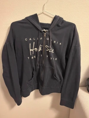 Mörkblå hoodie från Hollister - Mörkblå hoodie från Hollister med dragkedja, huva och fickor framtill. Tryckt text på bröstet med 'California Hollister Twenty Two'. Tillverkad i mjukt bomullsmaterial, perfekt för en avslappnad stil.