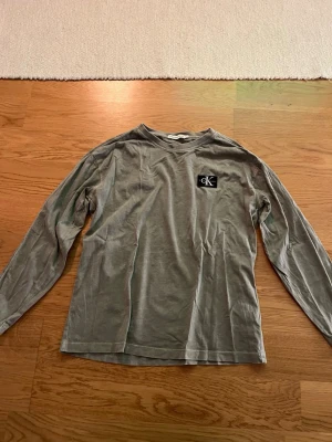 Grå långärmad tröja från Calvin Klein - Snygg grå långärmad tröja från Calvin Klein med CK-logga på bröstet. Tillverkad i mjukt bomullsmaterial och har en avslappnad passform. Perfekt till jeans eller kjol för en enkel och stilren look.